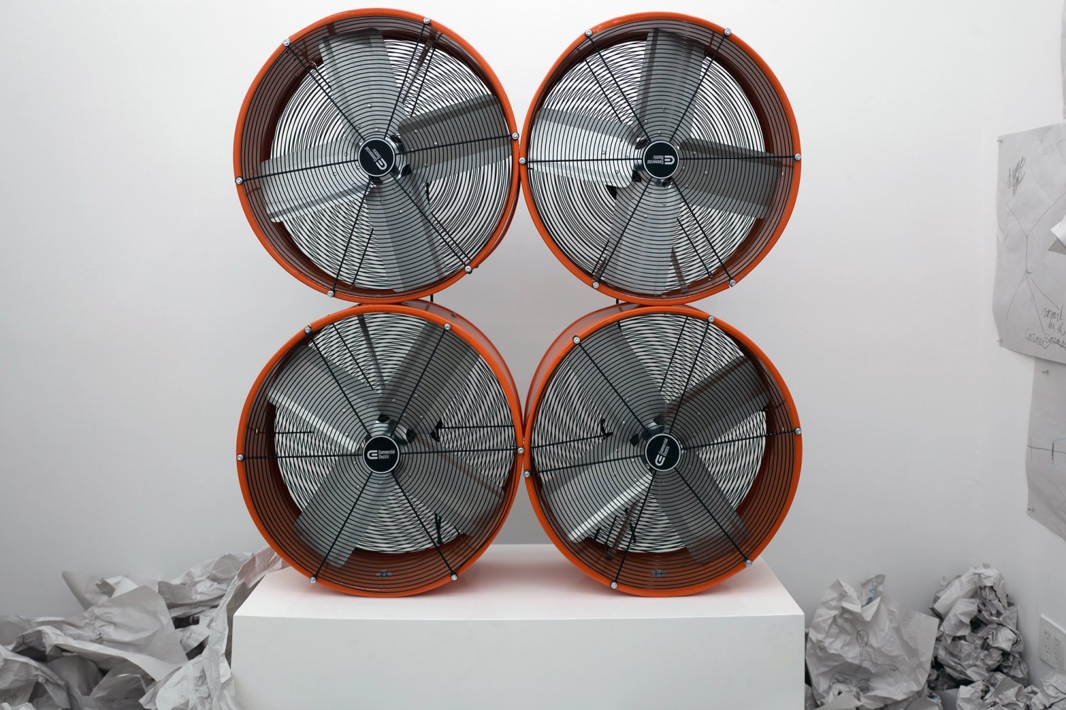 Four orange industrial fans stacked in a square above piles of discarded paper.