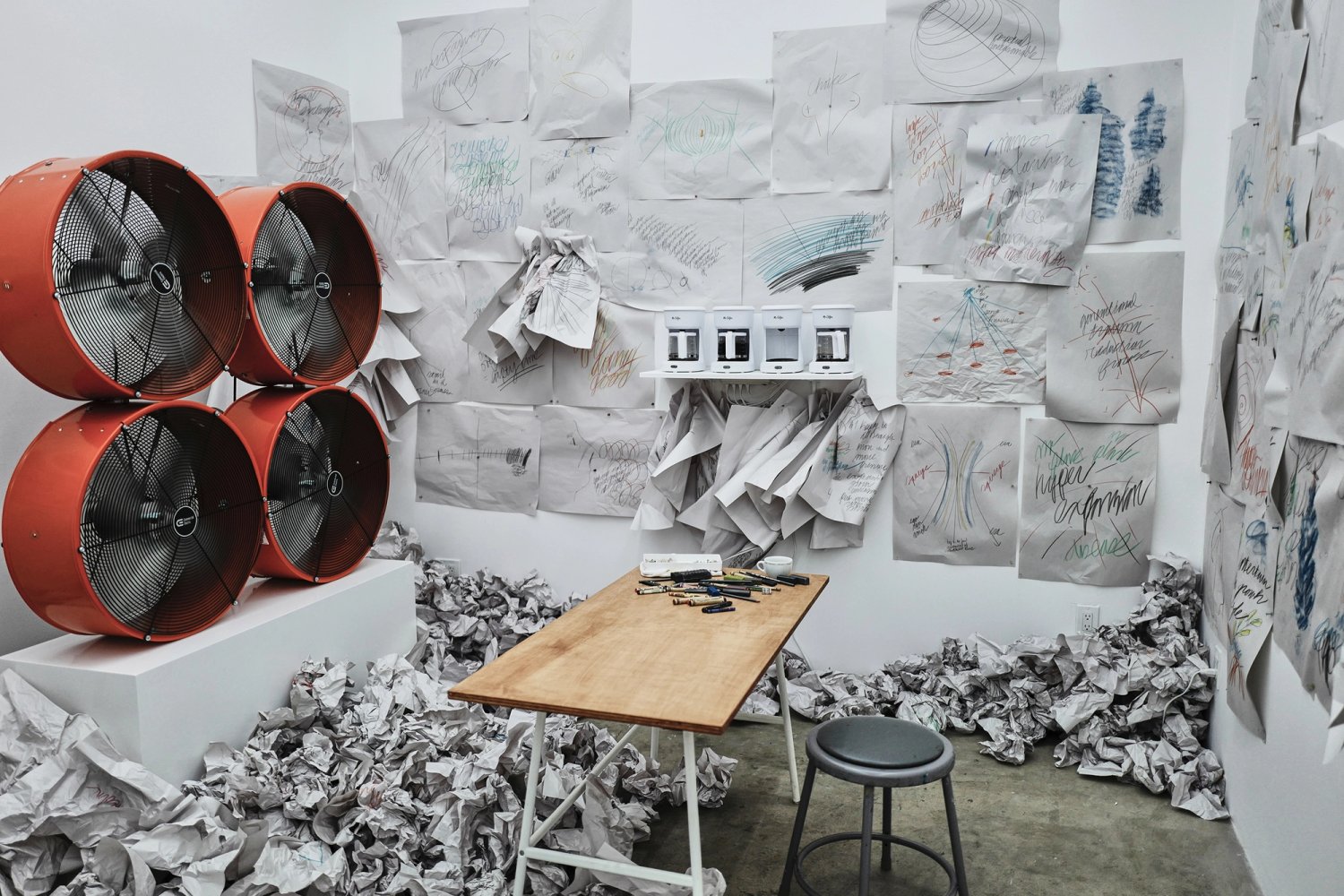 Four industrial fans face a drawing desk in a room covered with drawings and crumpled paper; four coffee machines sit on a shelf.