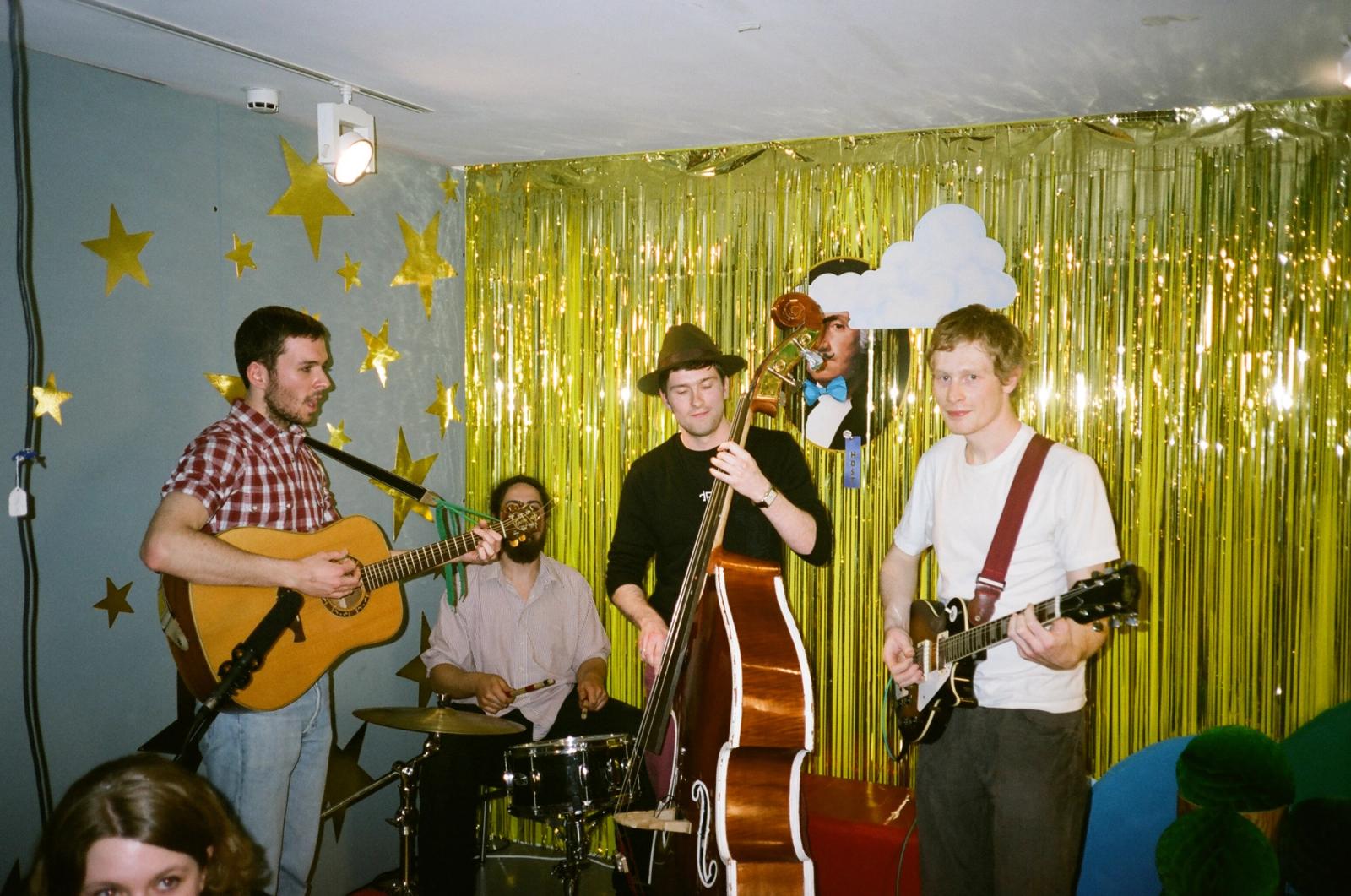 A live band performs with guitars, double bass, and drums against the talk show’s gold-curtain backdrop.