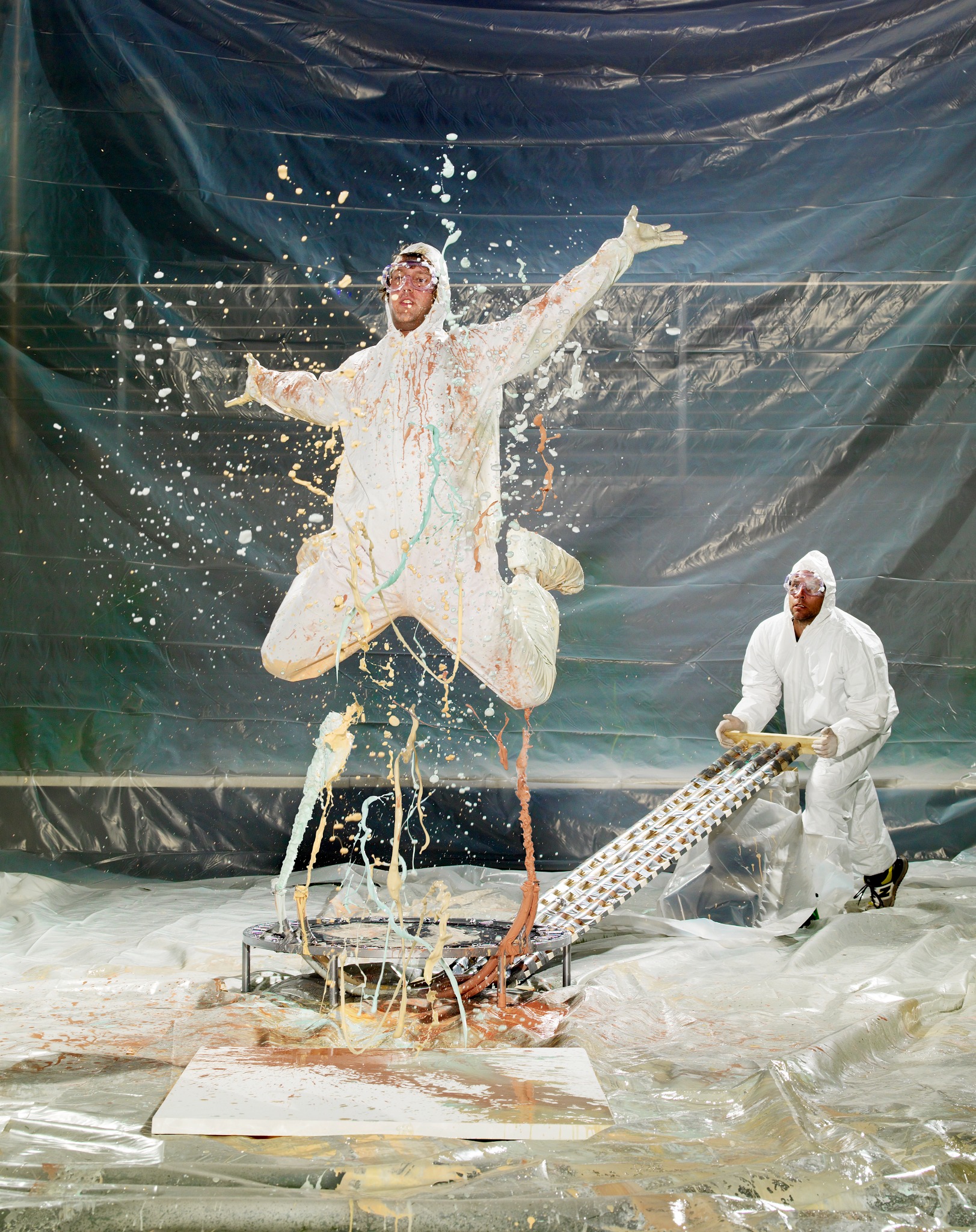 Marc leaps above a trampoline through airborne paint while a collaborator operates the painting apparatus.